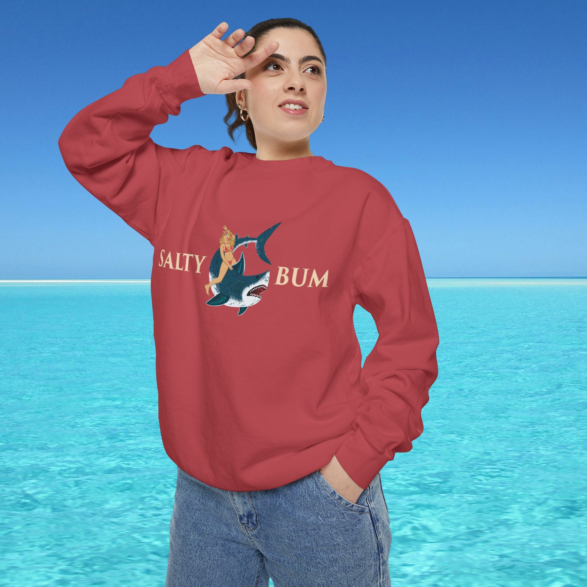 Person wearing a red sweatshirt with a graphic design and text, standing in front of a blue ocean and sky.
