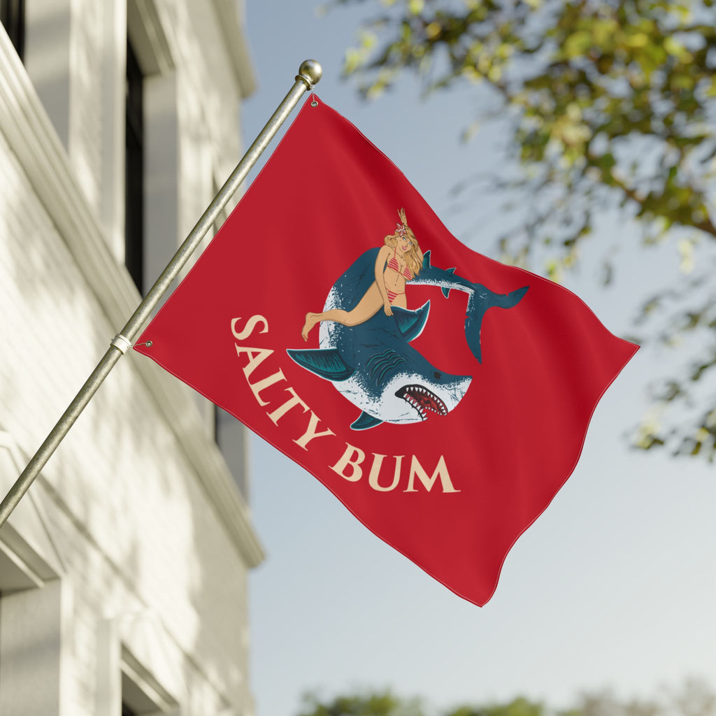 Red flag with a shark and woman design and 'Salty Bum' text, waving in front of a building.