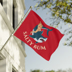 Red flag with a shark and woman design and 'Salty Bum' text, waving in front of a building.