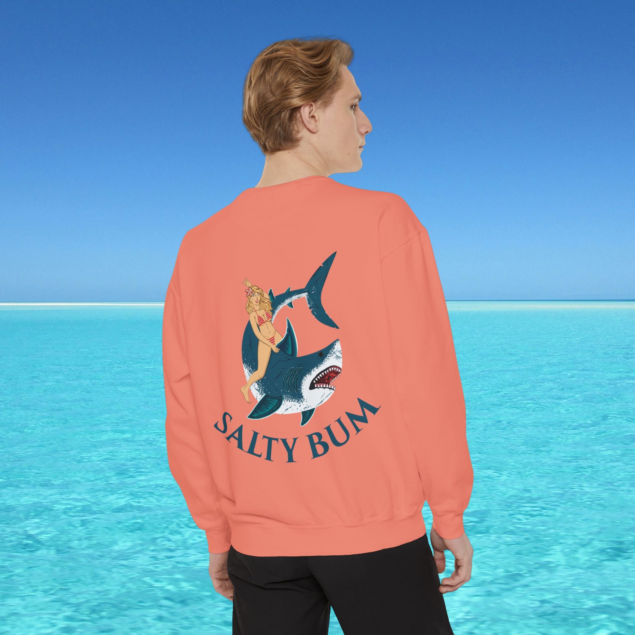 Person wearing a coral sweatshirt with a shark graphic and 'Salty Bum' text, standing in front of a blue ocean.