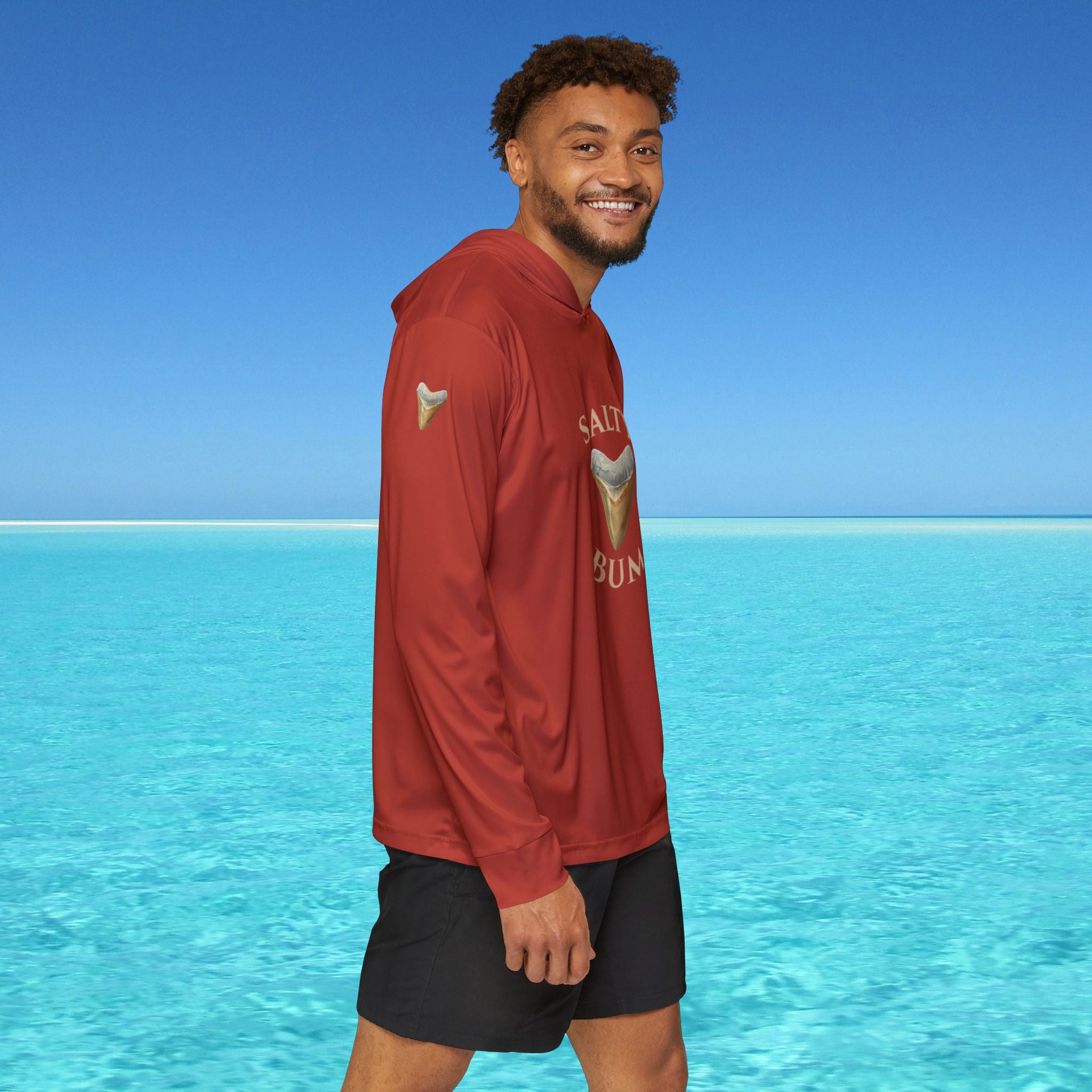 Man wearing a red hoodie with a logo standing in front of clear blue water and sky.