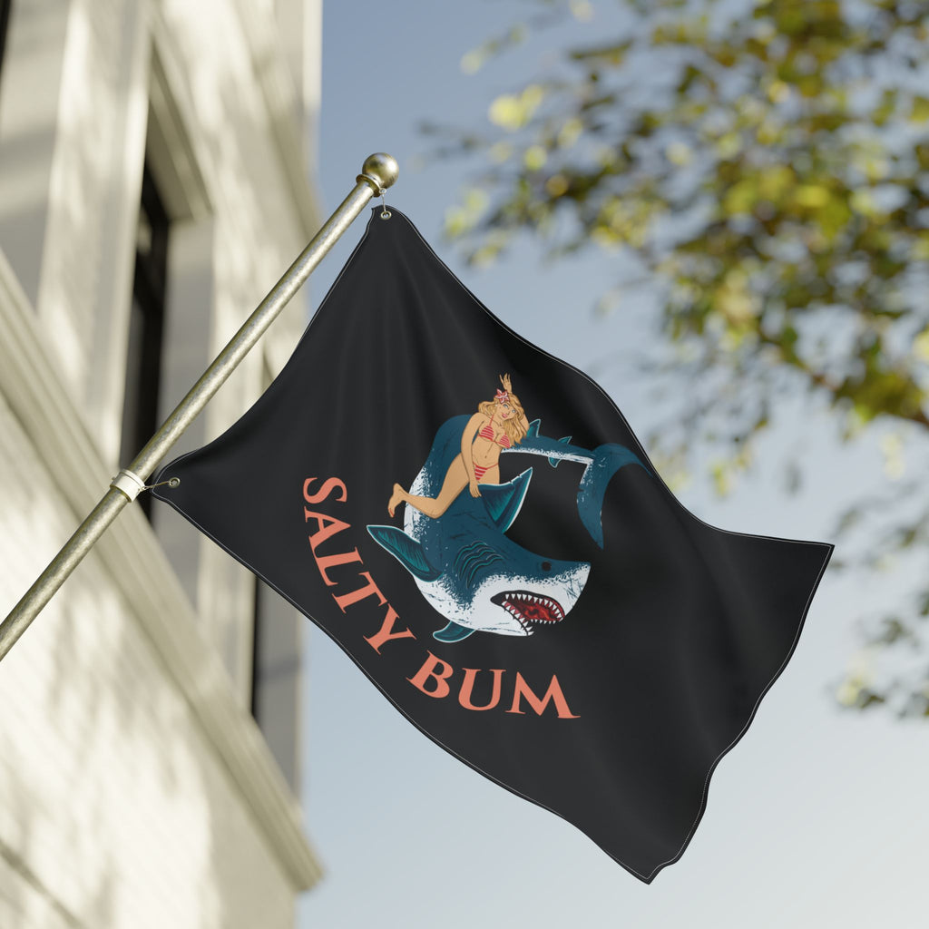 Flag with a shark and woman design and 'Salty Bum' text, waving against a building and tree.