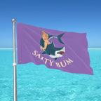 Flag with a mermaid and shark design on a purple background, waving in front of a clear blue sky and ocean.