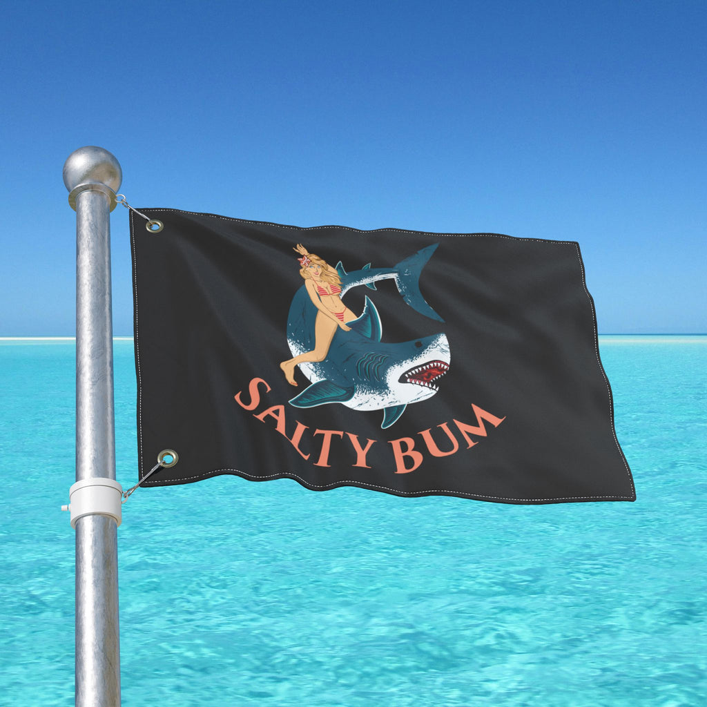 Flag with a graphic design of a person on a shark and 'Salty Bum' text, waving in front of a clear blue sky and ocean.