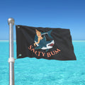 Flag with a graphic design of a person on a shark and 'Salty Bum' text, waving in front of a clear blue sky and ocean.