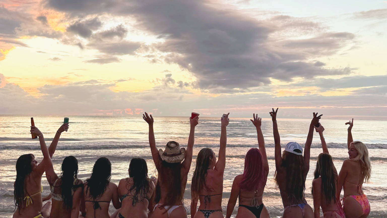 Group of women in bikinis raising their hands on a beach at sunset.